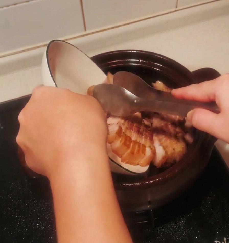 Searing pork belly pieces being poured from a white bowl back into the dark clay pot.