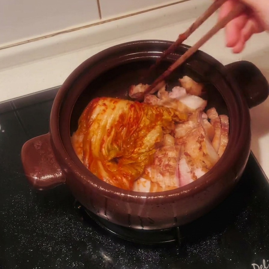 A whole head of seasoned red kimchi being placed into the clay pot over sliced pork belly using wooden chopsticks.