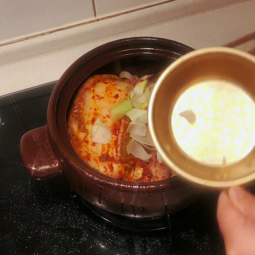 A gold metal bowl pouring sliced garlic and large green scallion segments onto the kimchi in the pot.
