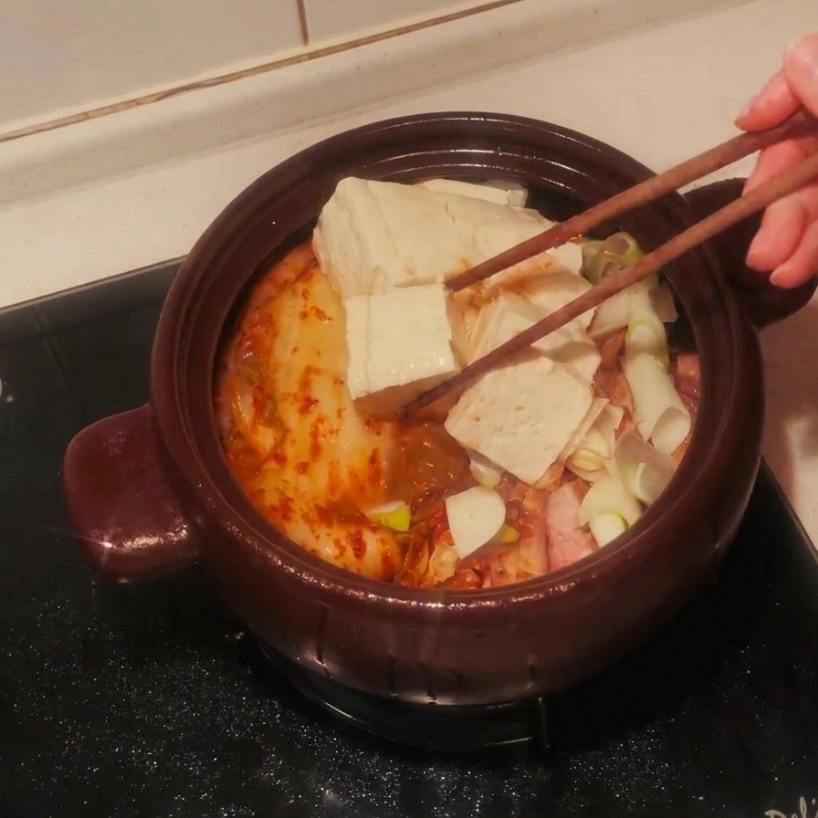Slices of white tofu being layered over the stew ingredients in a clay pot with chopsticks.