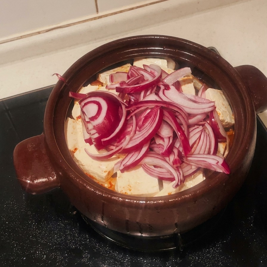 A pile of sliced red onions covering the layer of white tofu in the clay pot.
