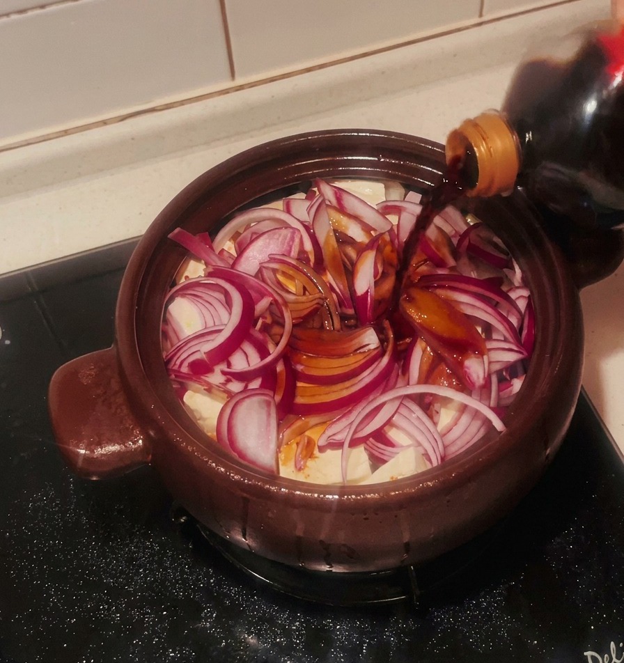 Dark soy sauce being poured from a bottle onto sliced red onions and tofu blocks inside a brown clay pot.