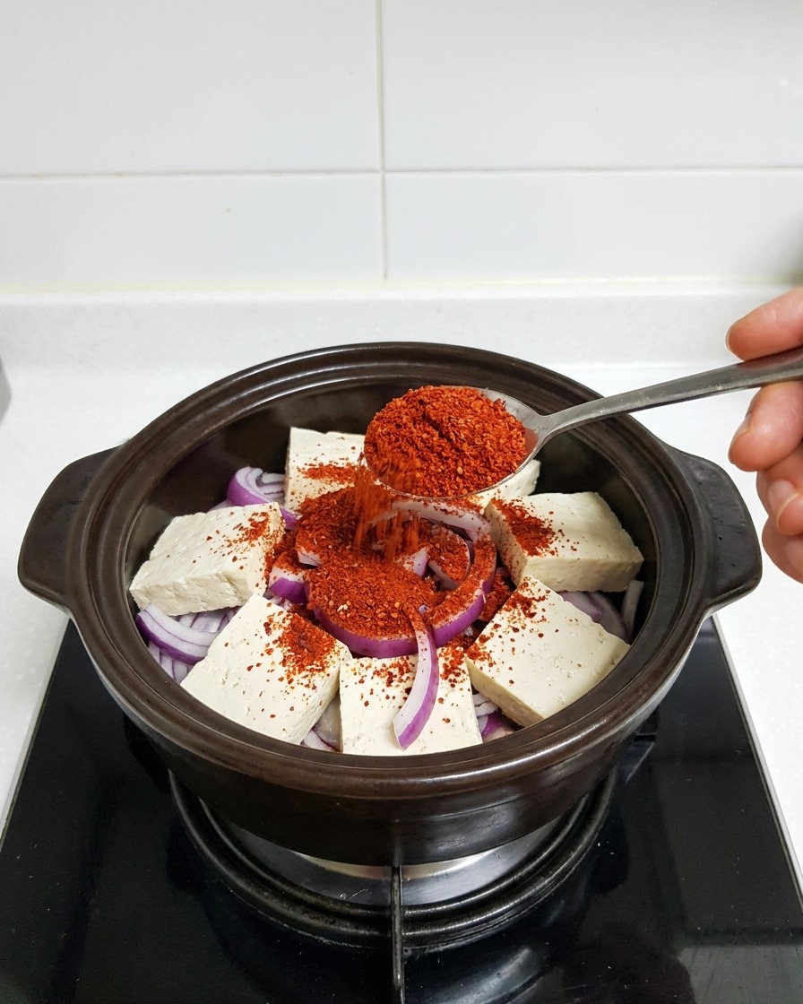 A metal spoon scooping bright red fine chili powder onto tofu and onions in a clay pot.