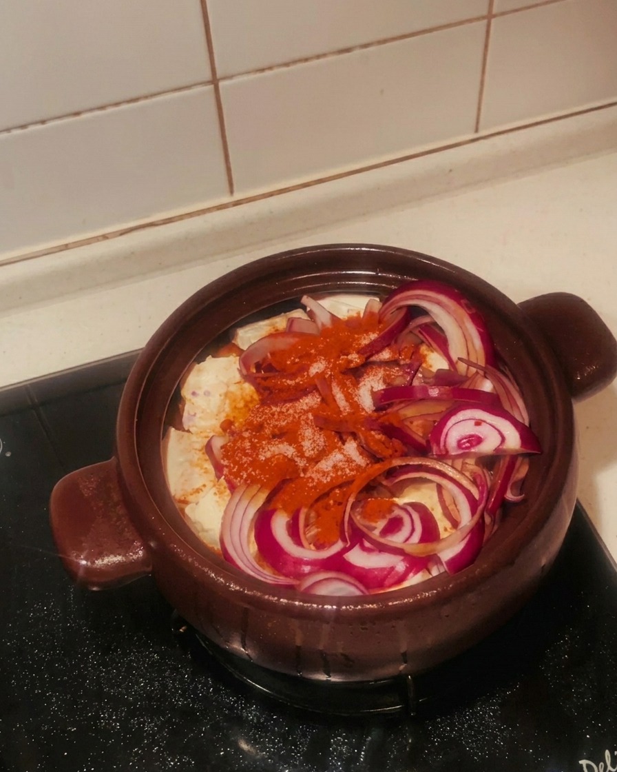 A hand sprinkling white granulated sugar over sliced red onions and chili powder in a brown clay pot.