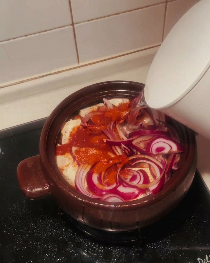 Clear water being poured from a white kettle into a clay pot filled with kimchi, tofu, red onions, and spices.