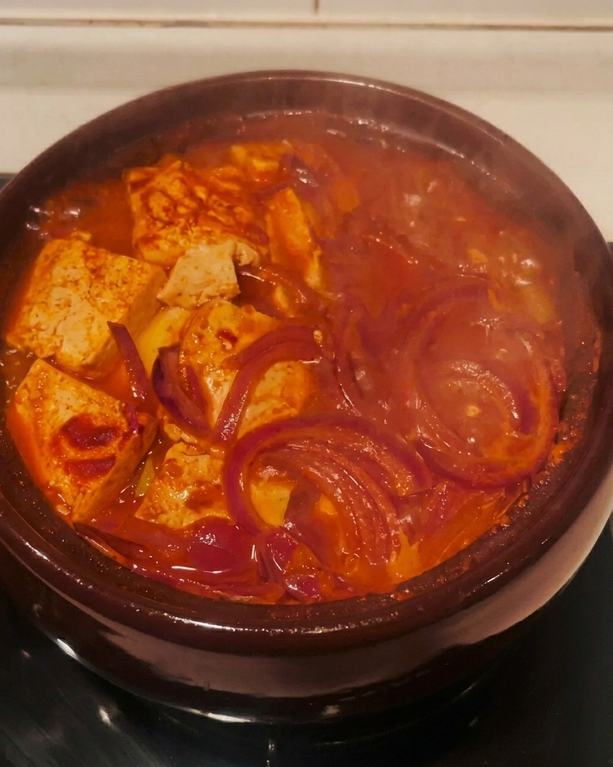 The red kimchi stew bubbling actively in a brown clay pot on a stovetop burner.