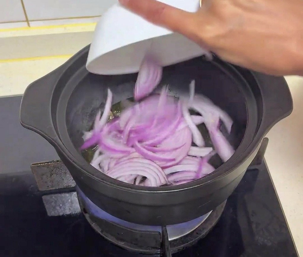 A hand pouring sliced red onions into a black pot containing heated oil and sliced shiitake mushrooms.