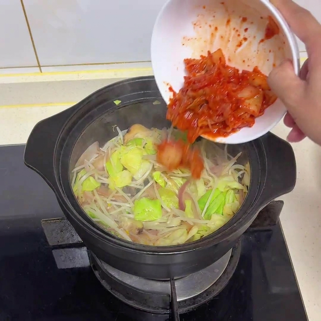 Bright red chopped kimchi being poured from a white bowl into a pot of sautéed vegetables.