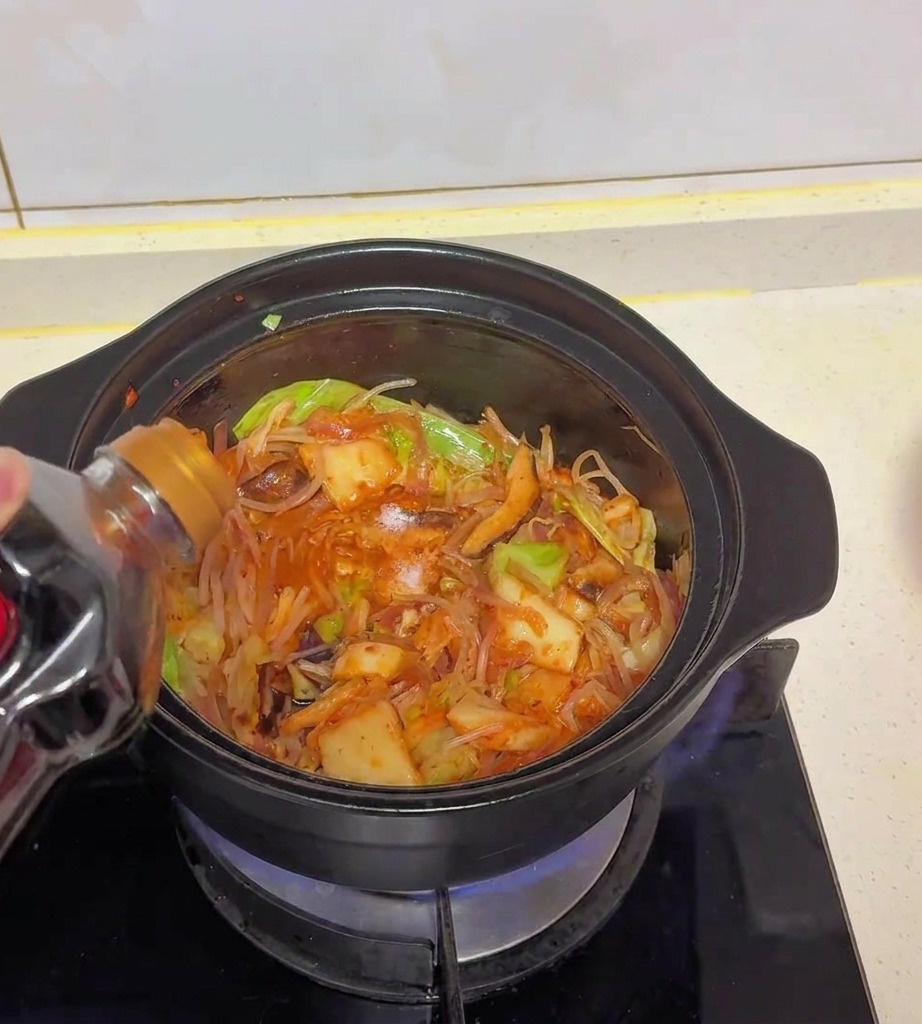 Light soy sauce being poured from a dark plastic bottle into a pot filled with sautéed cabbage, kimchi, and mushrooms.