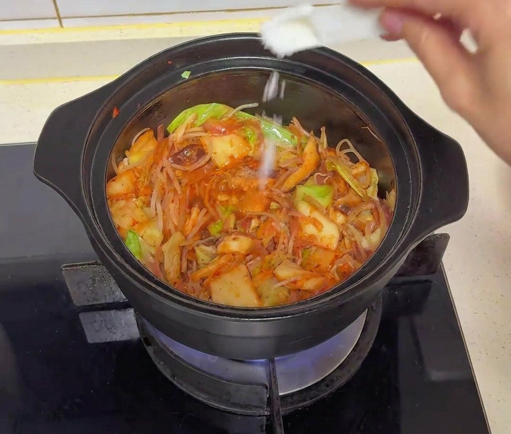 A hand using a small white spoon to sprinkle granulated sugar over the cooking vegetables in a black pot.