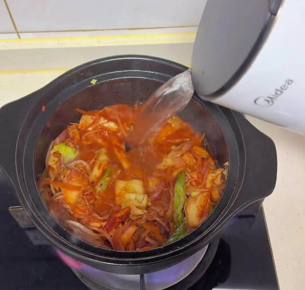 Clear hot water being poured from a white electric kettle into a black pot filled with sautéed kimchi and vegetables.
