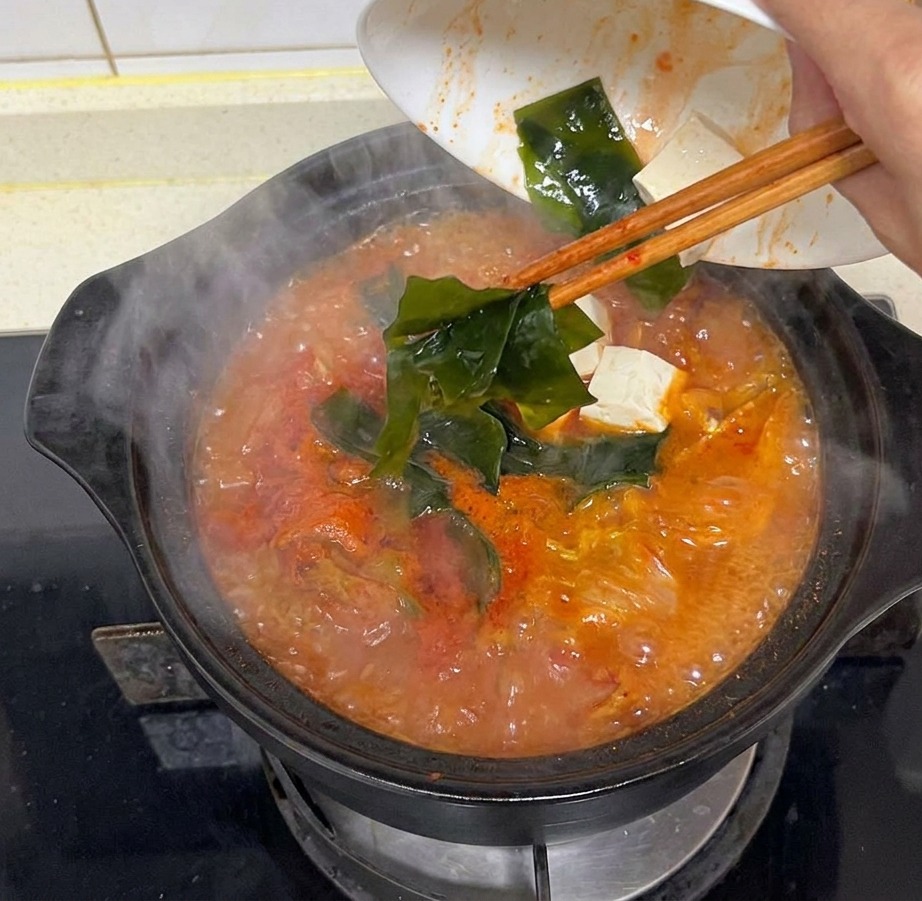 A hand pouring pieces of dark green kelp from a white bowl into a steaming pot of vibrant red kimchi soup bubbling on the stove.