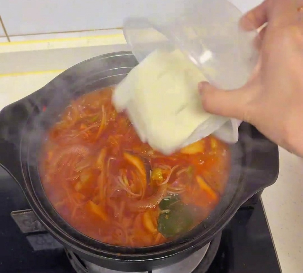 Fresh white tofu cubes being poured from a clear plastic container into a pot of boiling kimchi soup.