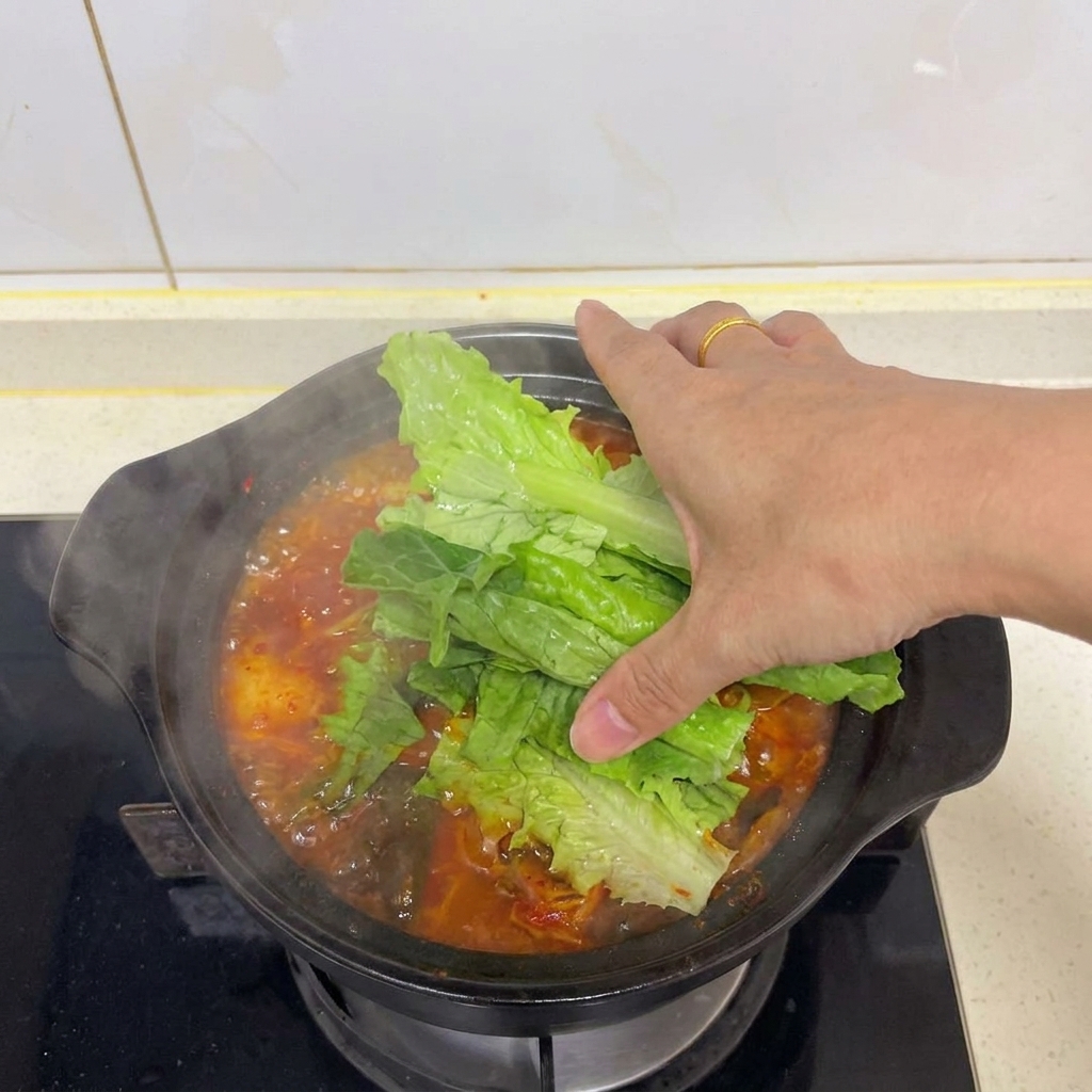 A hand placing vibrant green lettuce leaves over a steaming pot of red kimchi tofu soup.