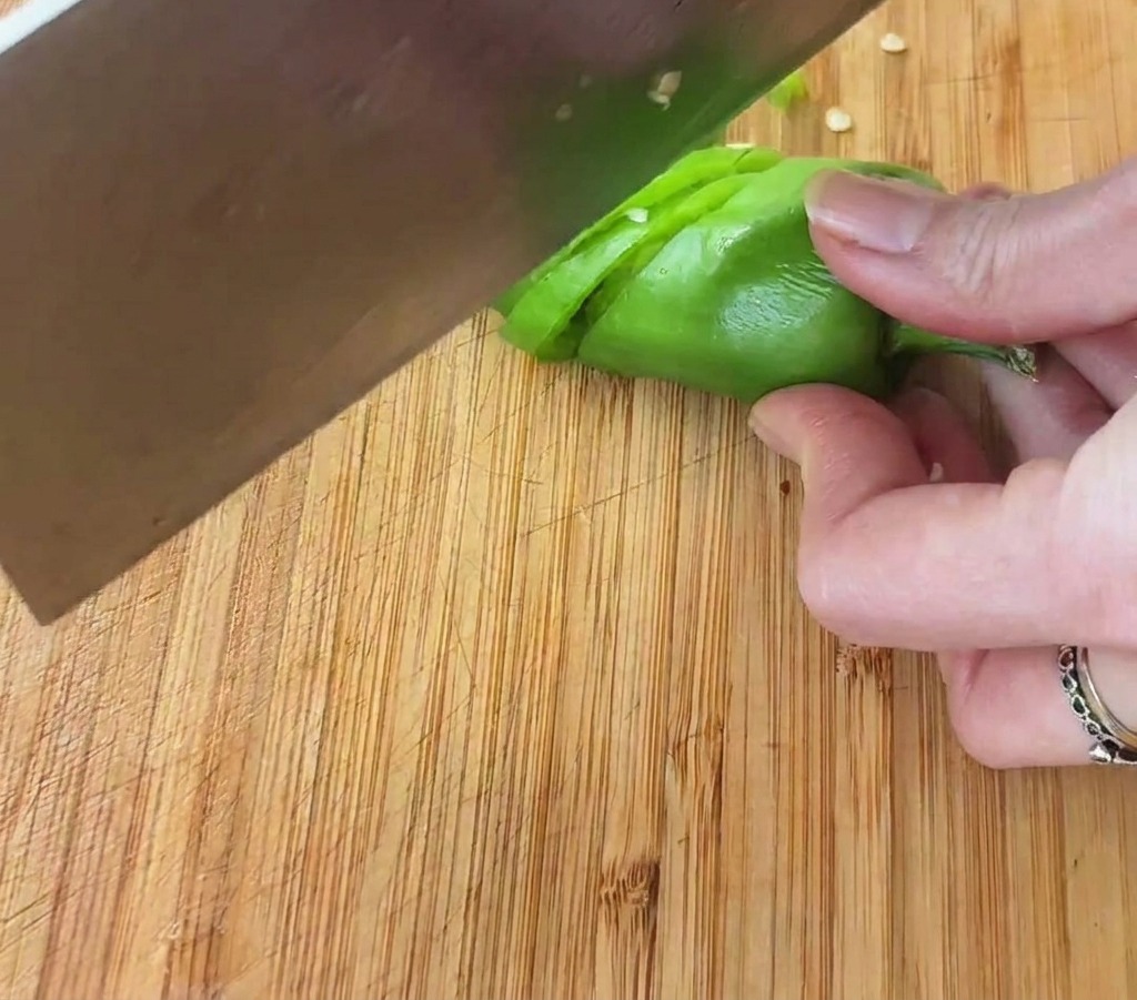 A close-up shot of a hand using a large metal cleaver to slice a bright green chili pepper into small rings on a wooden board.