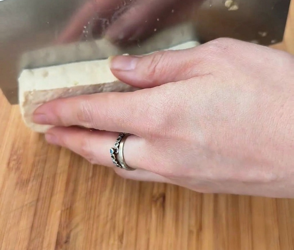A large rectangular block of white tofu being sliced into cubes with a wide metal cleaver on a wooden surface.