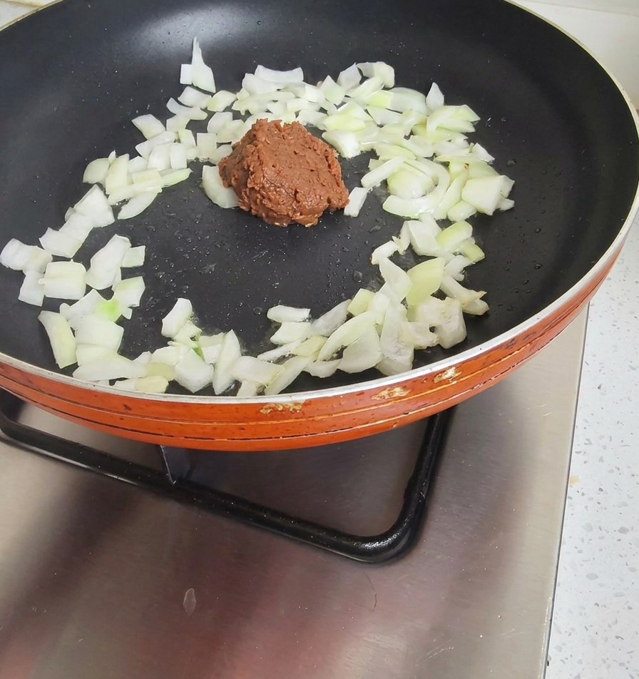 A dollop of thick, dark brown soybean paste added to a pan of partially sautéed white onions.