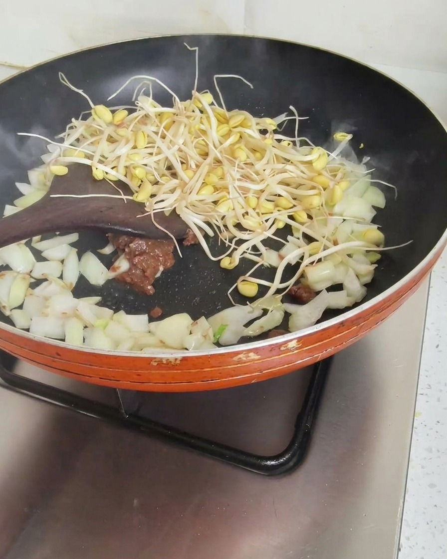 A wooden spatula stirring fresh bean sprouts, diced onions, and a scoop of brown soybean paste in an orange frying pan.
