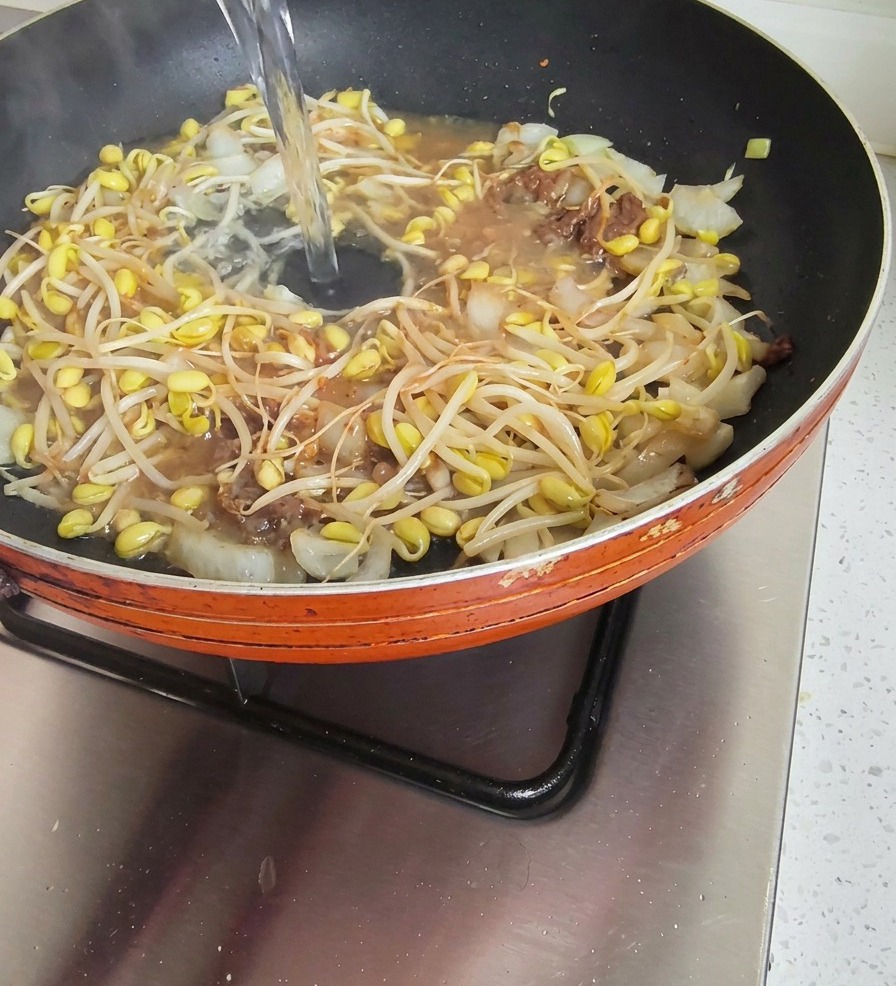 A stream of clear water being poured into a frying pan filled with bean sprouts and onions in a savory soybean paste base.
