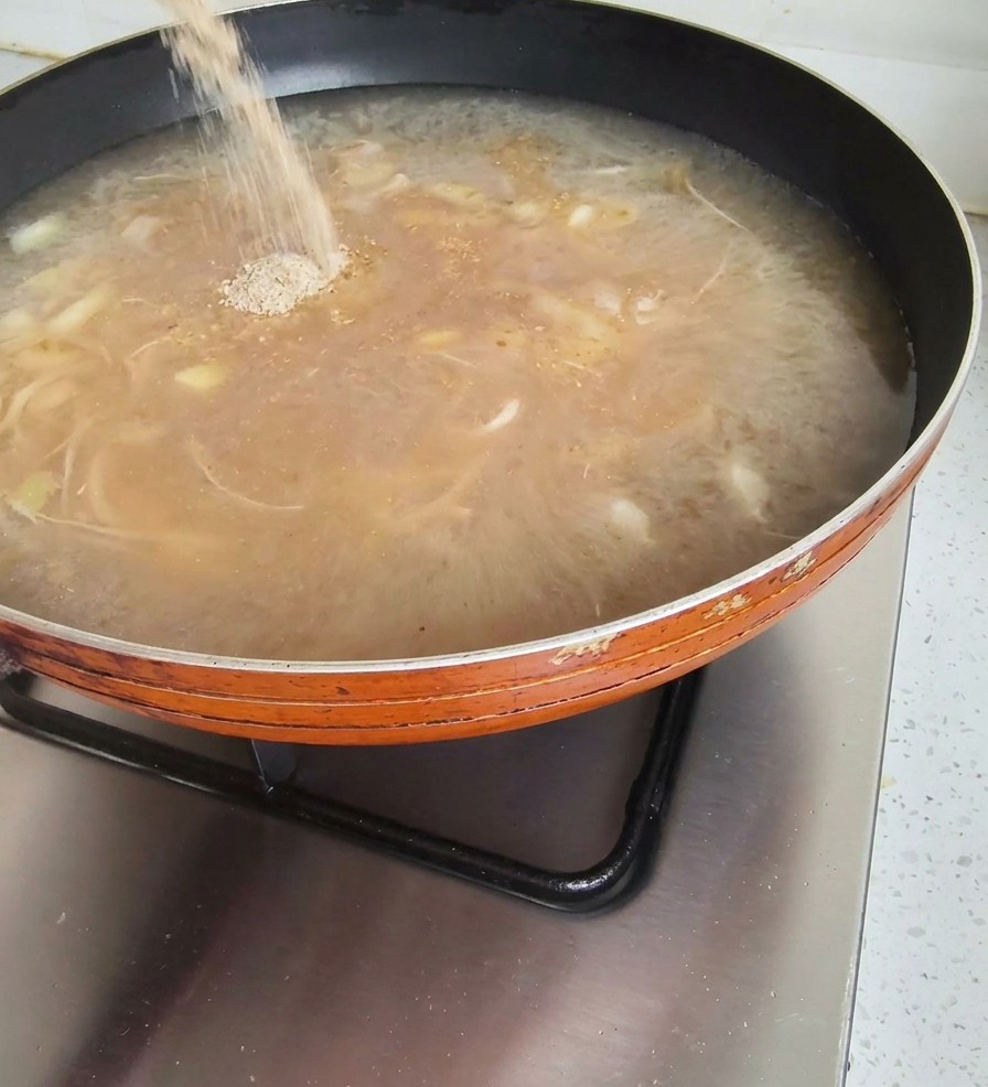 Seasoning powder being poured from a packet into a pan of bubbling brown soup broth on a stovetop.
