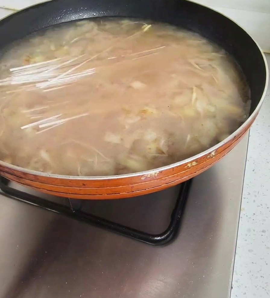 A handful of dry, translucent noodles being placed into a pan of simmering Korean soybean paste soup.