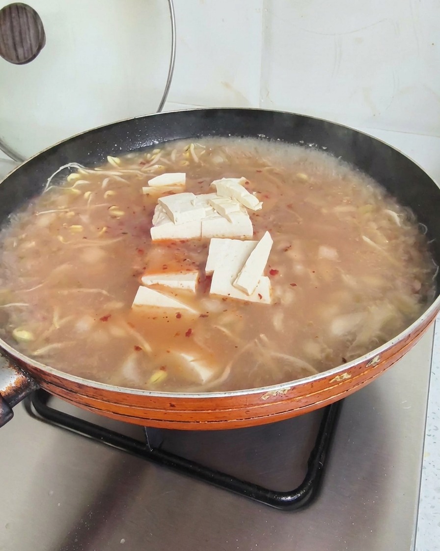 Tofu cubes resting on the surface of a simmering reddish soup in a large orange frying pan.