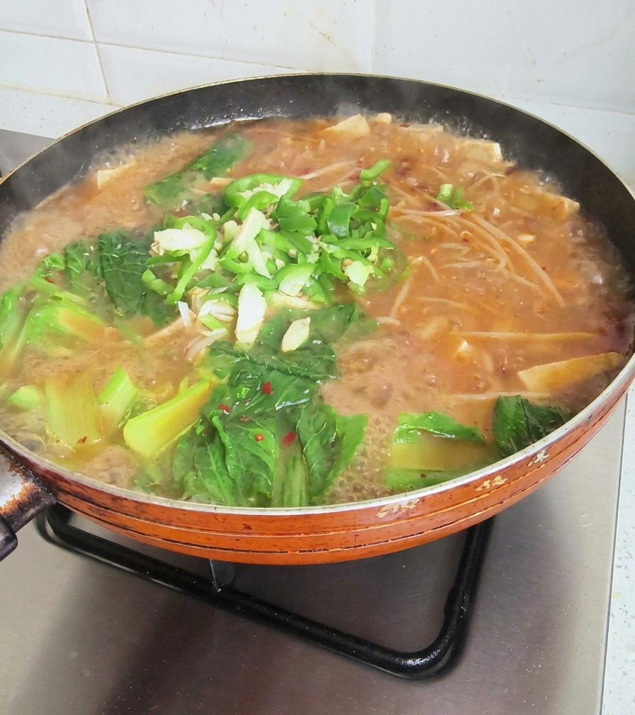 A bubbling pan of Korean soybean soup topped with fresh green leafy vegetables, sliced green chilies, and minced garlic.