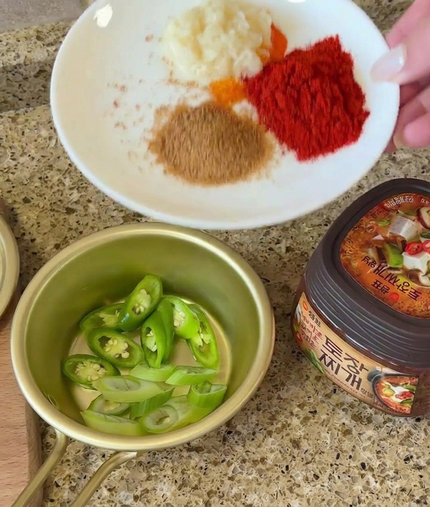 A hand holding a plate with portions of garlic and spices next to a bowl of sliced chilies and a jar of soybean paste.