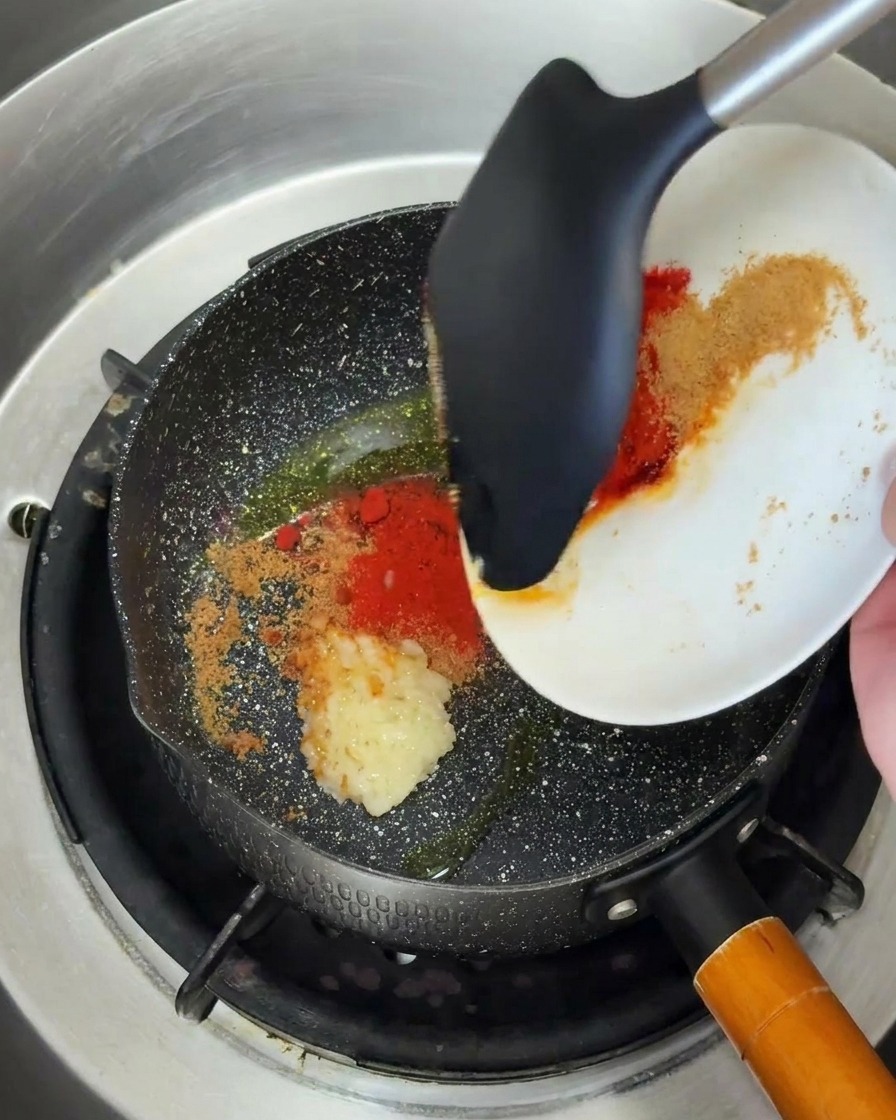 Minced garlic and red spice powder being scraped from a white plate into a hot pan with oil using a black spatula.