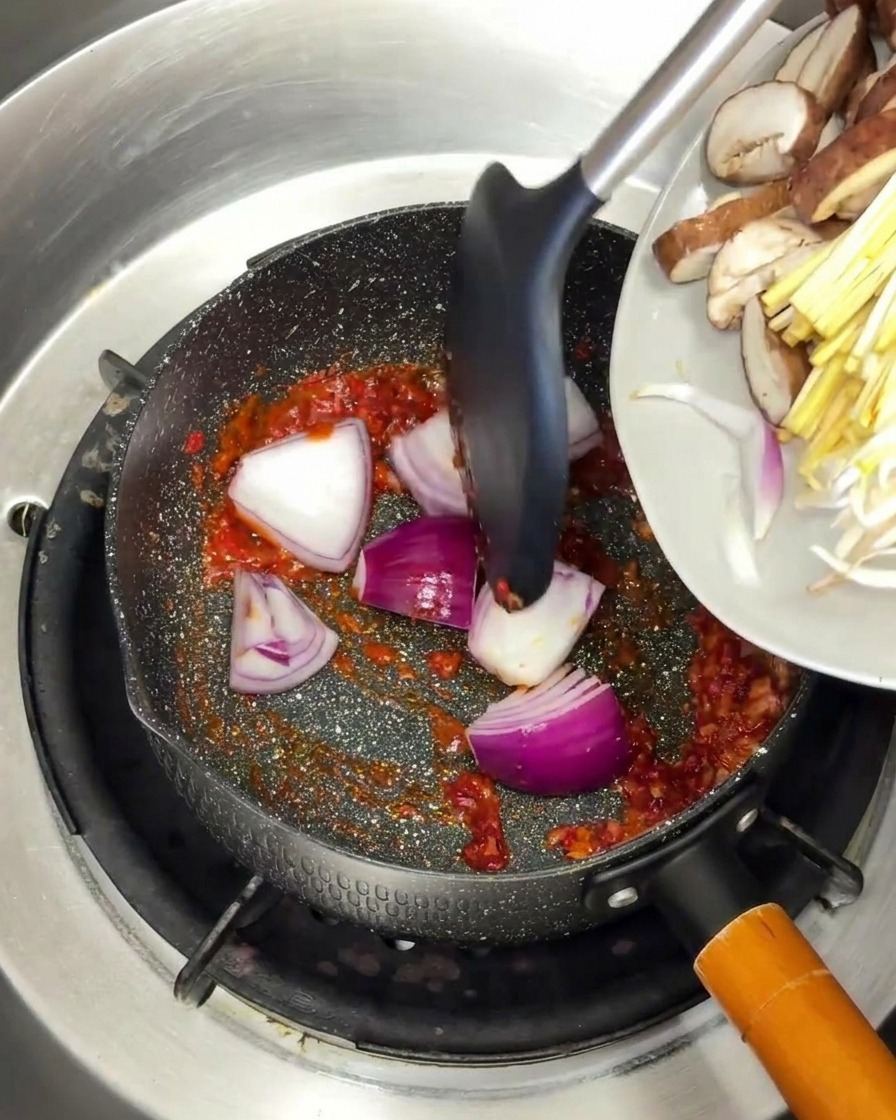 Red onion wedges and sliced mushrooms being added to a pan with a sizzling red spice base.