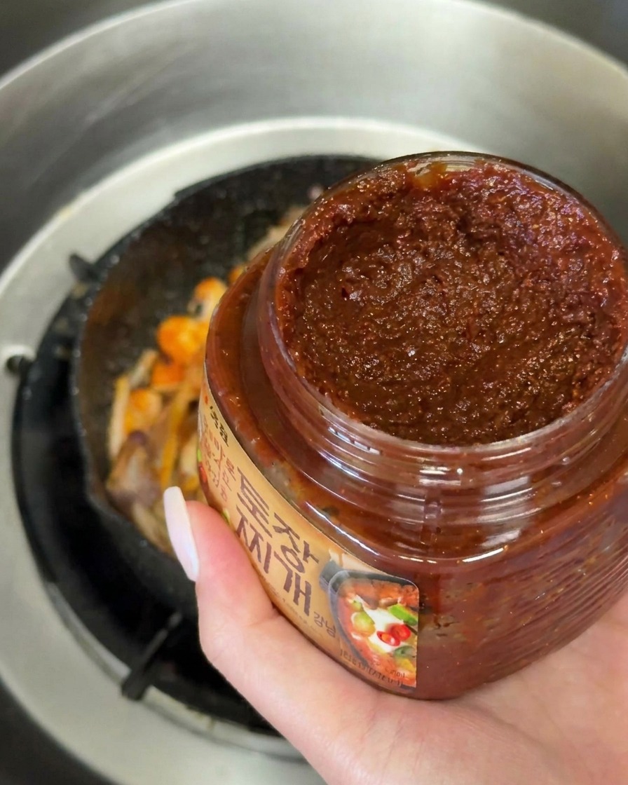 A hand holding a jar and a large spoonful of thick dark brown Korean soybean paste over a pan of sizzling vegetables.