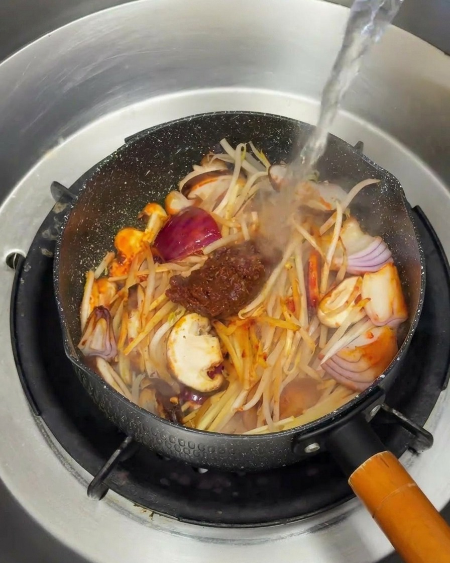 A stream of hot water being poured into a pan over sautéed vegetables and a dollop of dark brown soybean paste.