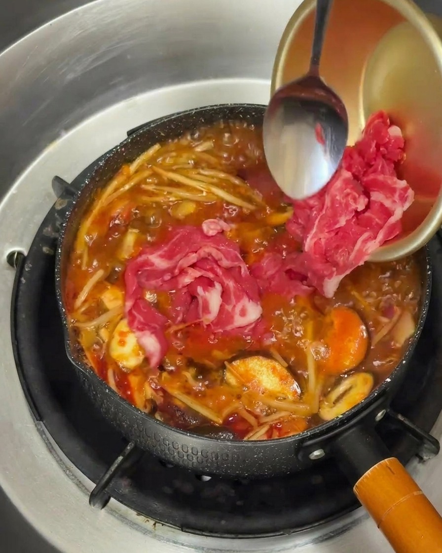 Marbled slices of raw beef being added from a gold colored bowl into a bubbling pot of vibrant red soup broth.
