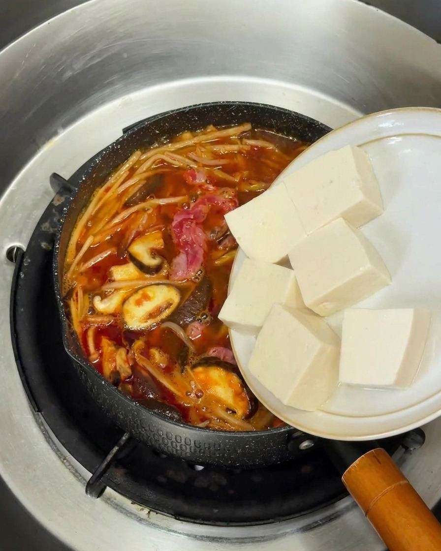 Fresh cubes of soft white tofu being moved from a white plate into a pot of bubbling Korean beef soup.