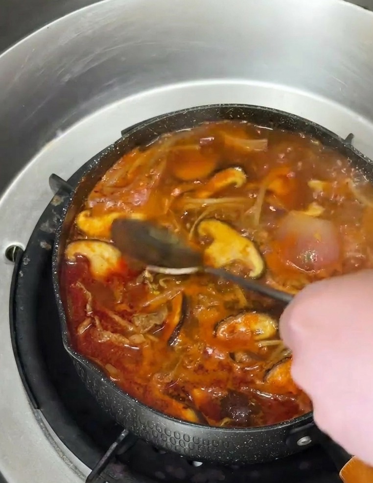 A metal spoon pouring a stream of translucent amber sesame oil into a bubbling pot of red, spicy Korean beef soup filled with mushrooms.
