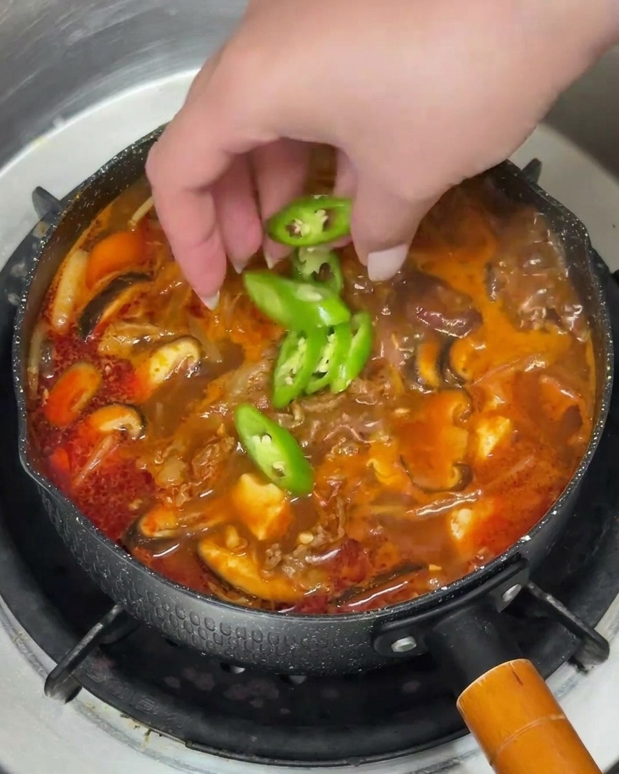 A hand scattering bright green chili rings and chopped scallions into a steaming pot of red soup.