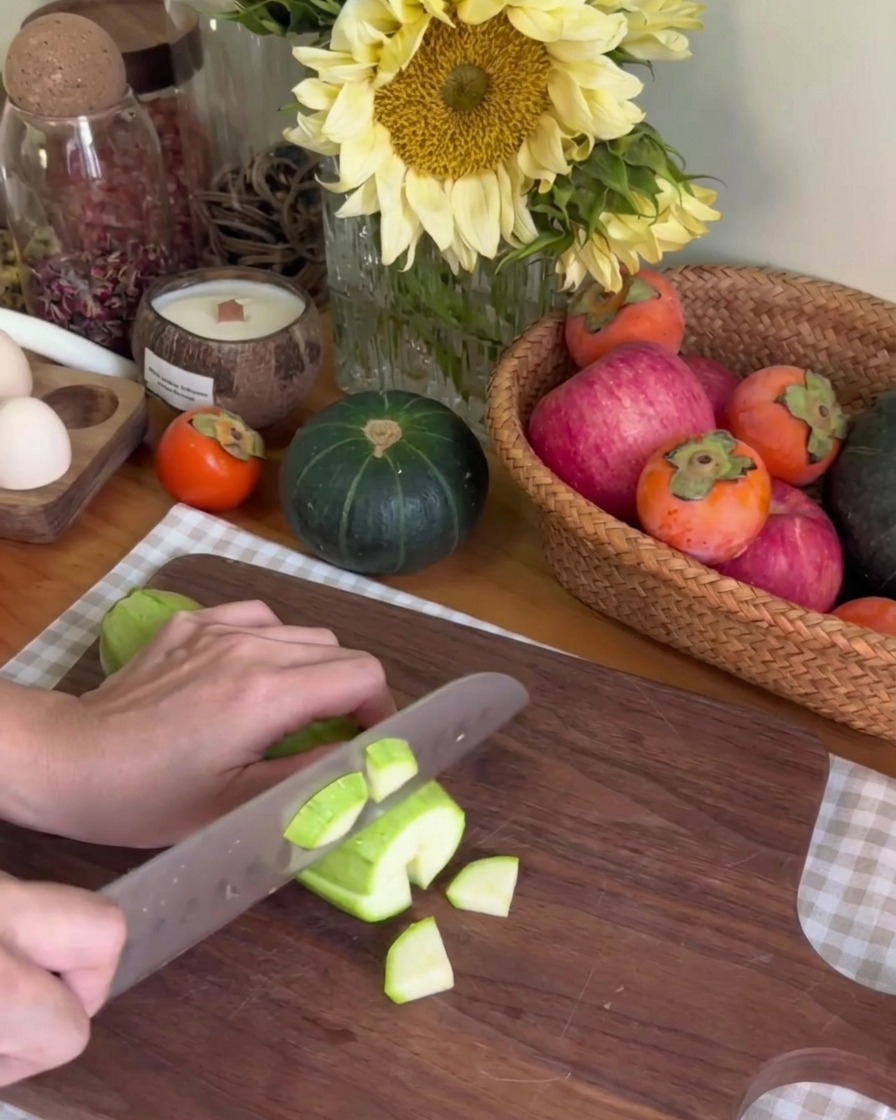 A person using a kitchen knife to slice a green zucchini into small pieces on a dark wood chopping board