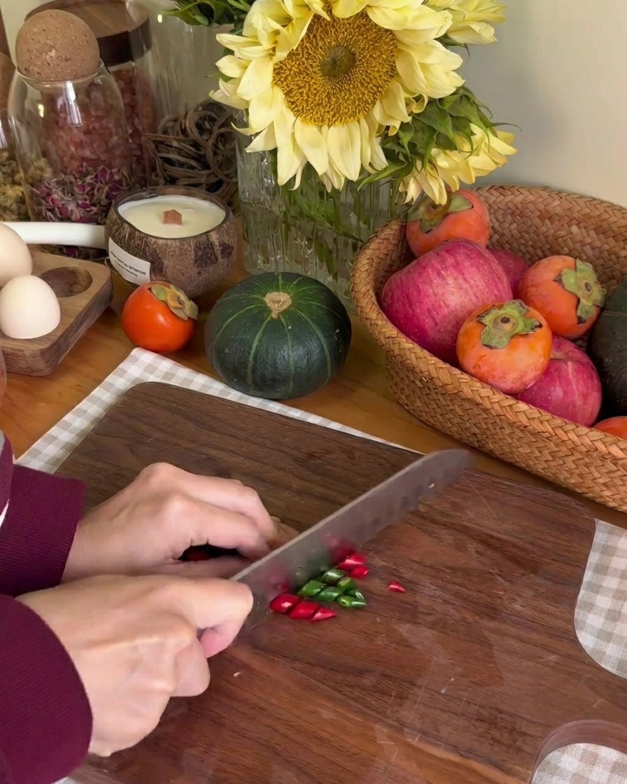 A person using a knife to slice small green and red chilies into thin rings on a wooden board.