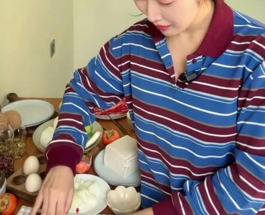 A person using a kitchen knife to finely mince fresh cloves of garlic on a wooden surface surrounded by other vegetables like onions and zucchini.