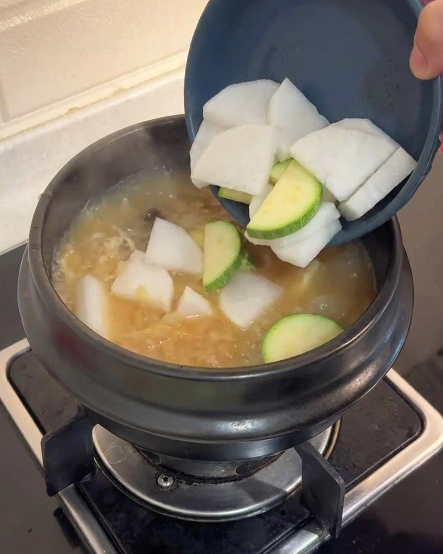 Slices of white radish and green zucchini being poured from a blue bowl into a boiling pot of brown broth.