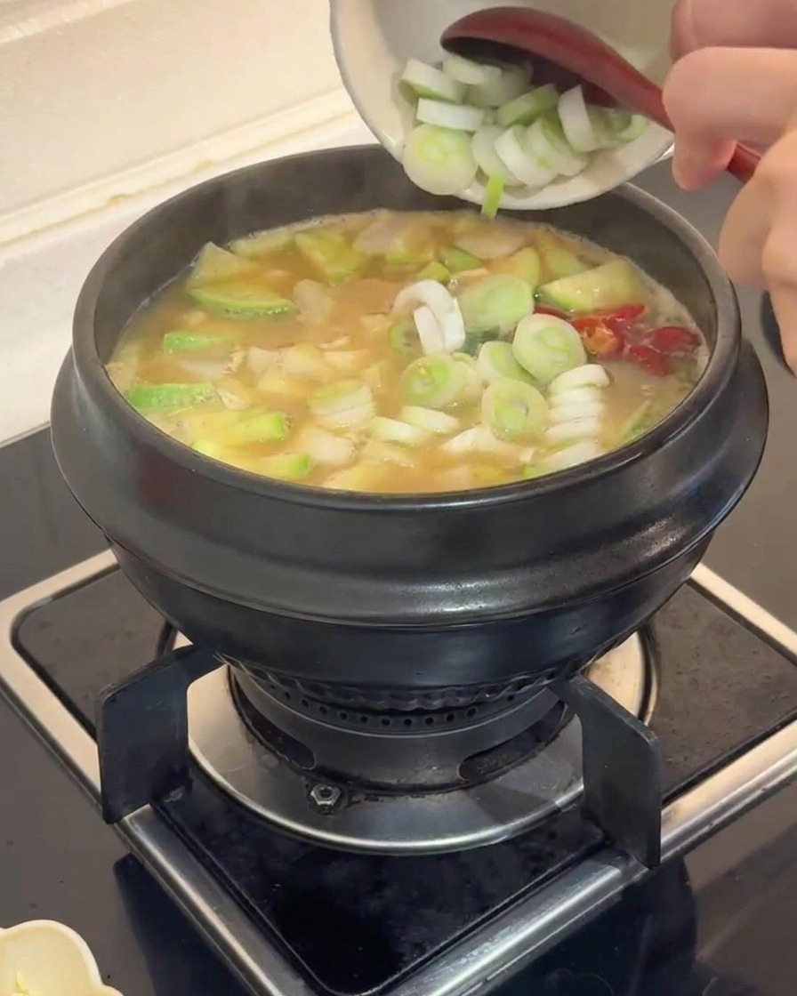 Sliced green leeks being added from a small white bowl into a bubbling stone pot of vegetable stew.