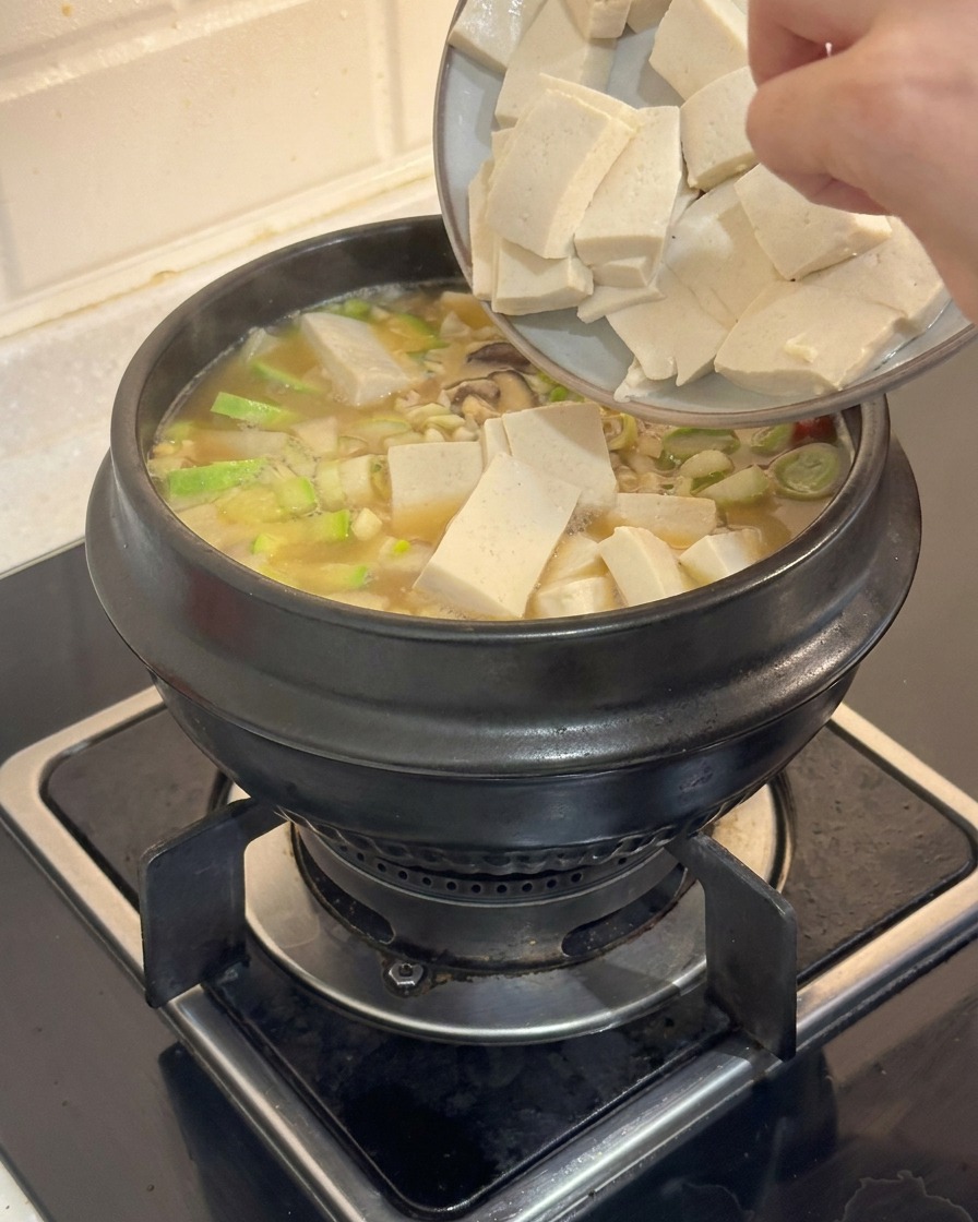 A hand sliding rectangular cubes of white tofu from a plate into a steaming pot of Korean stew.