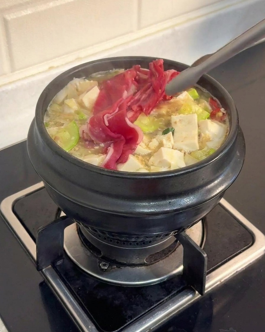 Metal tongs placing thin, marbled slices of raw beef into a pot filled with tofu and vegetables in a bubbling broth.