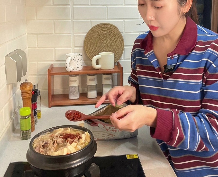 A wooden spoon adding bright red chili powder to a bubbling pot of Korean stew filled with beef slices, tofu, and zucchini.