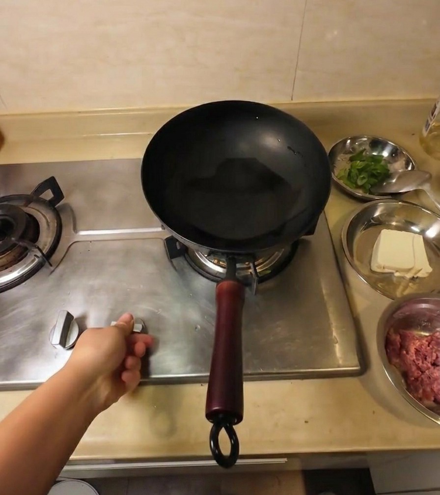 A hand adjusting the gas stove knob under a black wok that contains clear cooking oil.