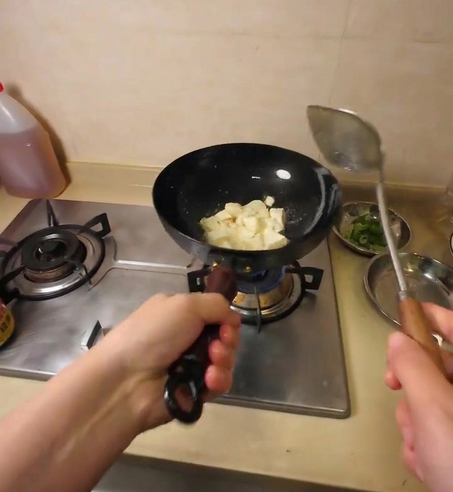 A metal spatula breaking a large block of tofu into smaller cubes while it sizzles in hot oil.