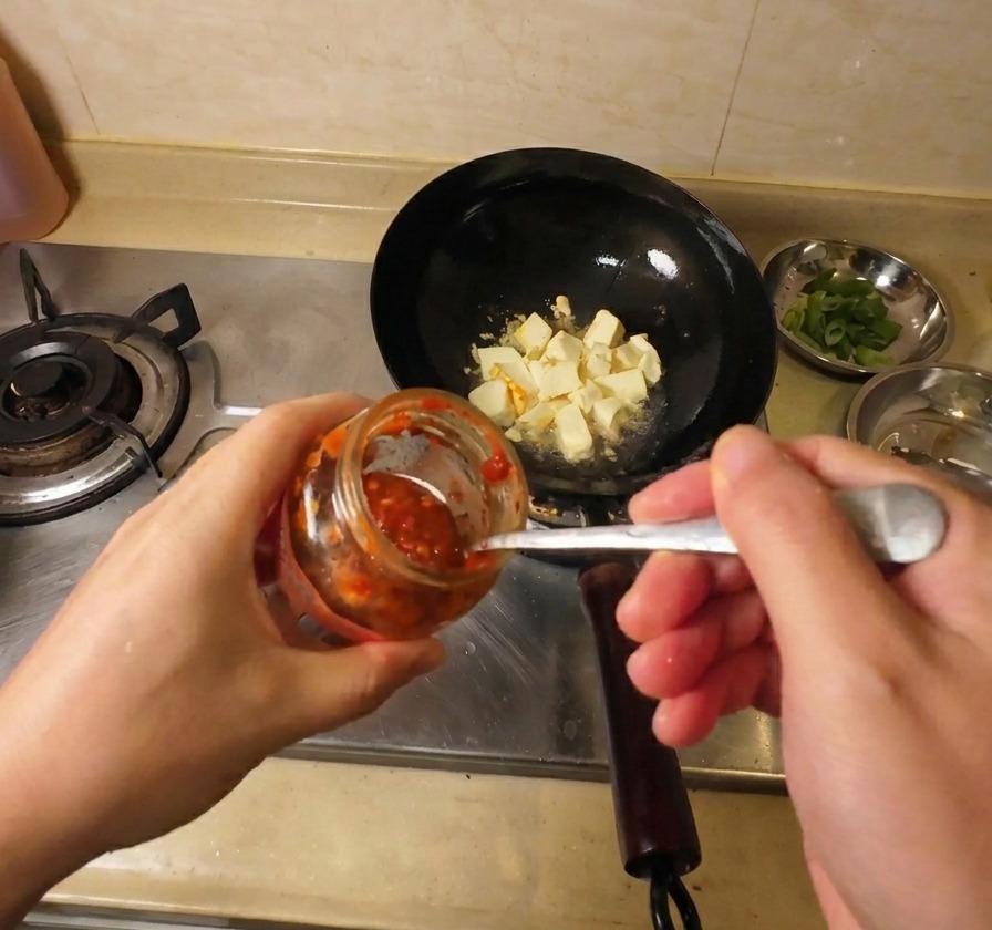 A close-up shot of a hand using a metal spoon to add vibrant red chili sauce from a glass jar into a black wok filled with golden-brown fried tofu pieces.