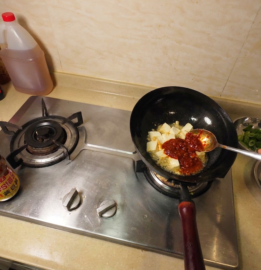 Dark brown soybean paste on a metal spoon being added to the wok alongside fried tofu and red chili sauce.