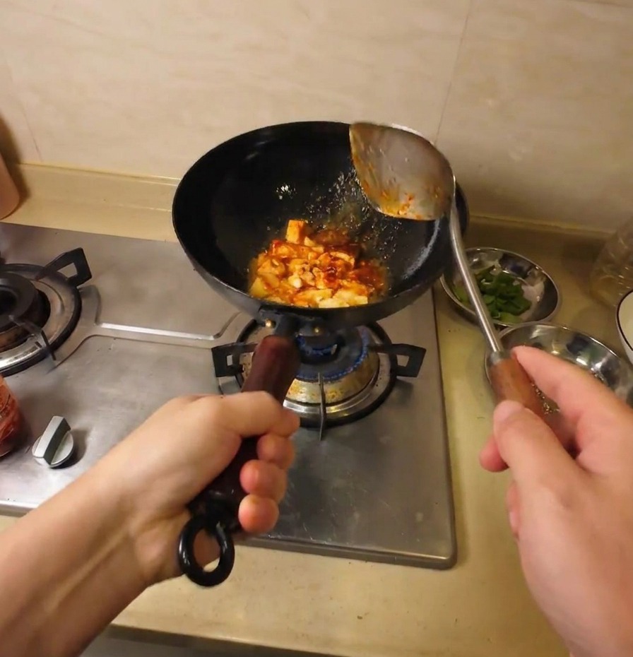 Tofu pieces being tossed and coated in a savory red sauce in a black wok.