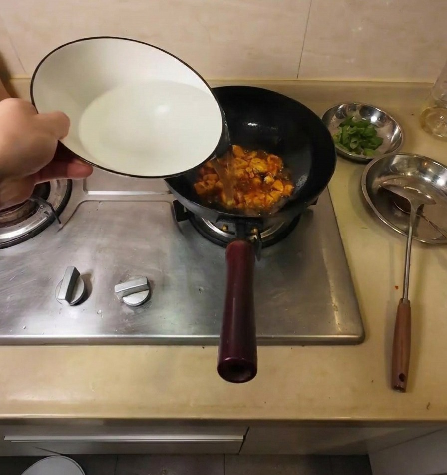 Hot water being poured from a white ceramic bowl into a black wok containing fried tofu and red seasonings.
